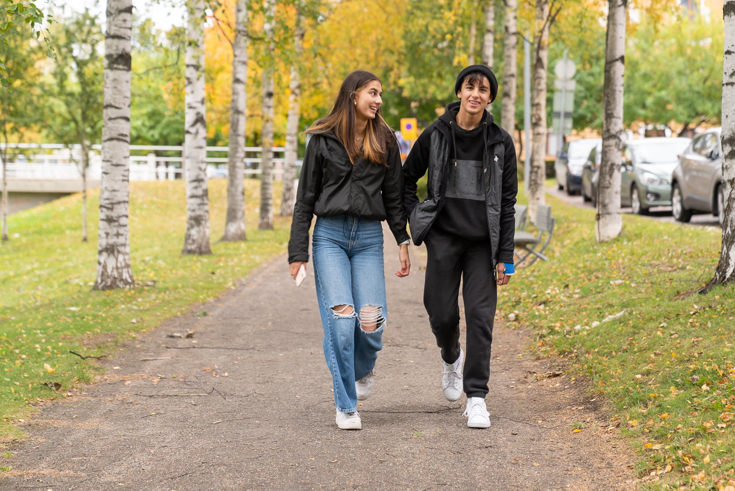 Kaksi hymyilevää nuorta kävelee rinnakkain kävelytiellä syksyisessä maisemassa. Taustalla koivuja, joiden alla keltaisia lehtiä.