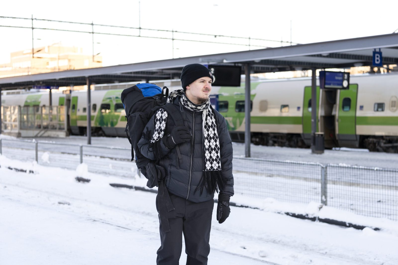 Nuori mies talvisessa maisemassa reppu selässään. Taustalla juna.