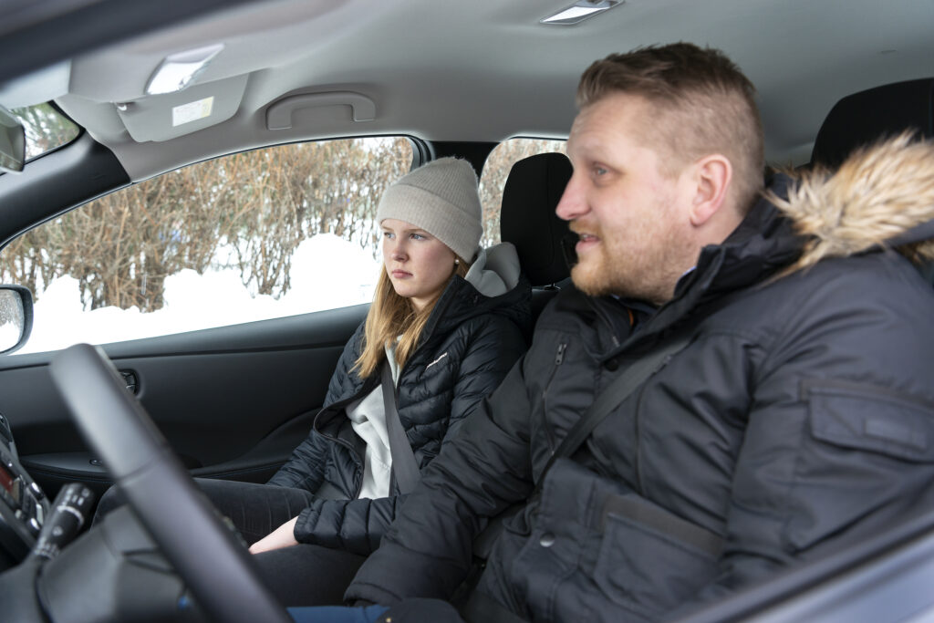 Autossa aikuinen mies kuljettajan paikalla ja nuori nainen pelkääjän paikalla. Molemmilla tarvitakki. Auton ikkunasta näkyy lunta.