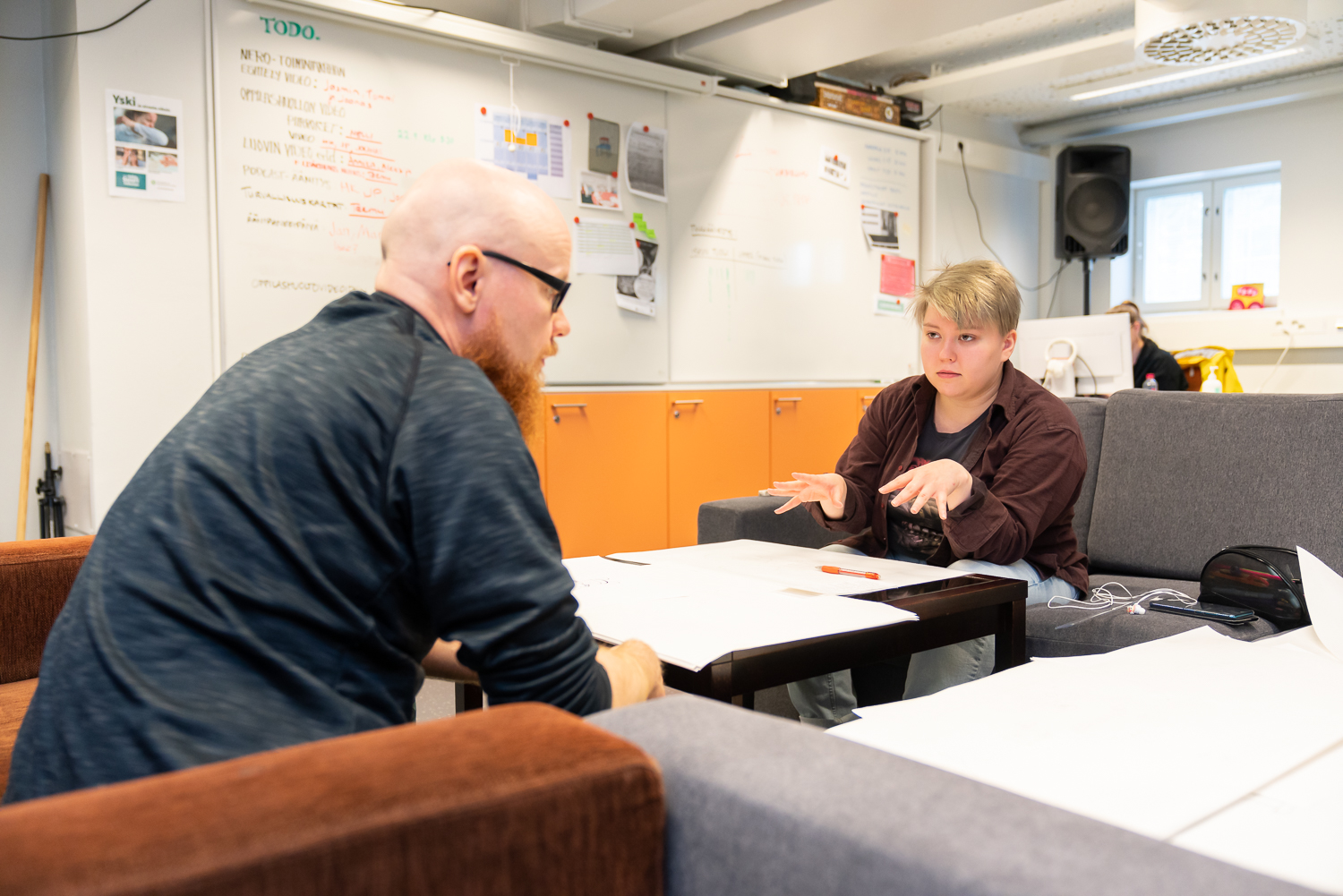 Kalju mies, jolla silmälasit ja parta, istuu vastapäätä nuorta. Nuori katsoo miestä kädet rinnan korkeudella. Nuoren ja miehen välissä pöytä, jossa papereita ja kynä.