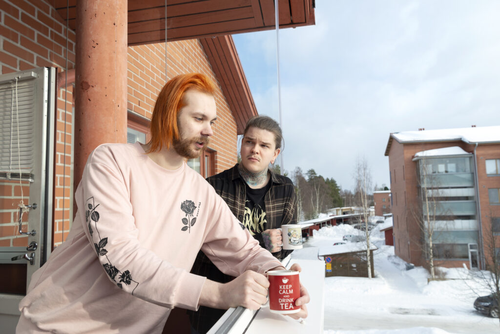 Kaksi nuorta miestä kerrostalon parvekkeella kupit kädessään. Toinen katsoo eteenpäin suu auki toisen katsoessa häntä. Toisen kupissa lukee Keep calm and drink tea. Alhaalla maassa lunta.