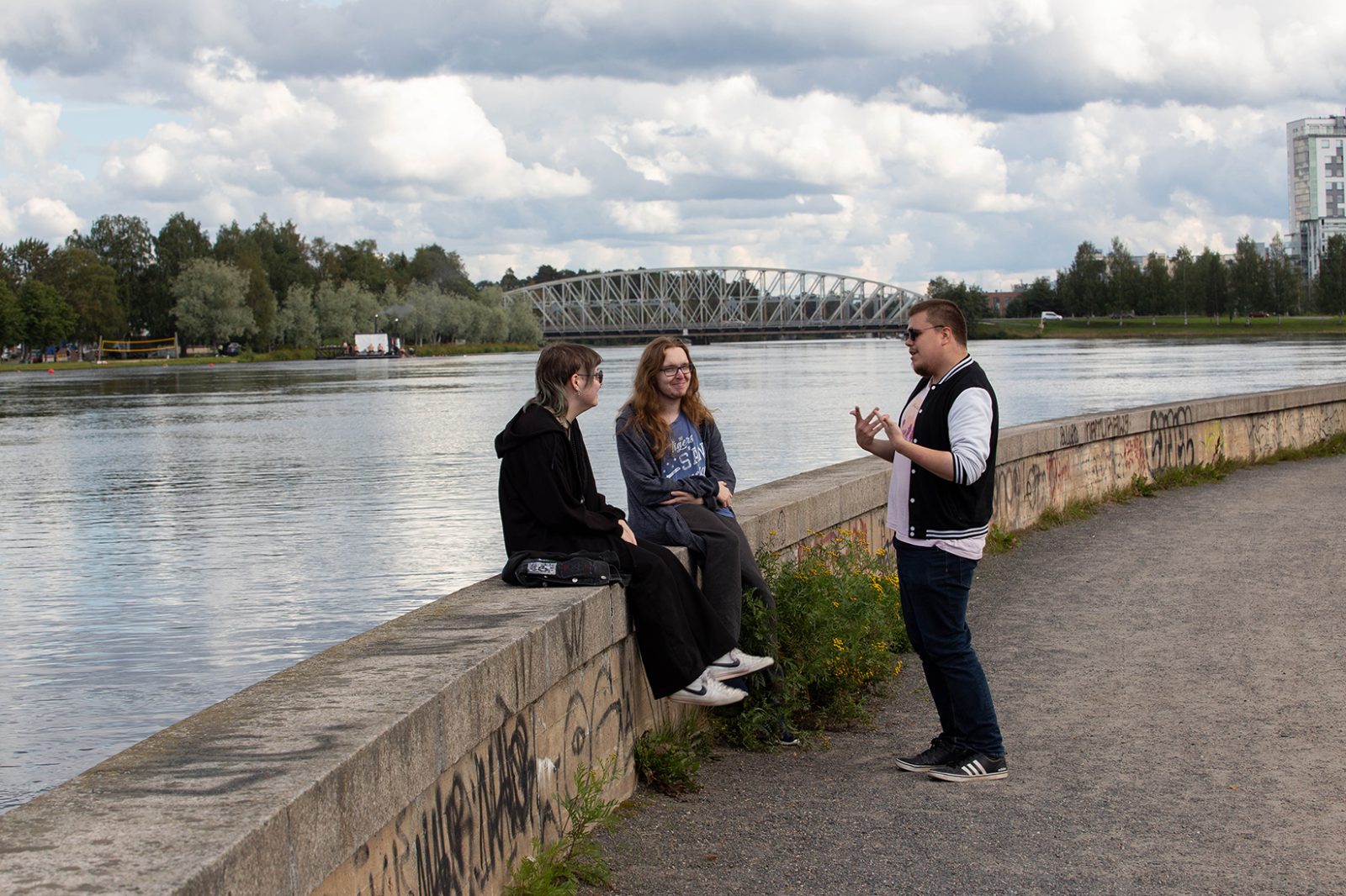 Kolme henkilöä veden äärellä, yksi seisoo ja kaksi istuu betonireunalla. Taustalla on silta, jossa on ainutlaatuinen geometrinen rakenne, sekä rakennuksia, jotka viittaavat kaupunkiympäristöön. Taivas on osittain pilvinen.