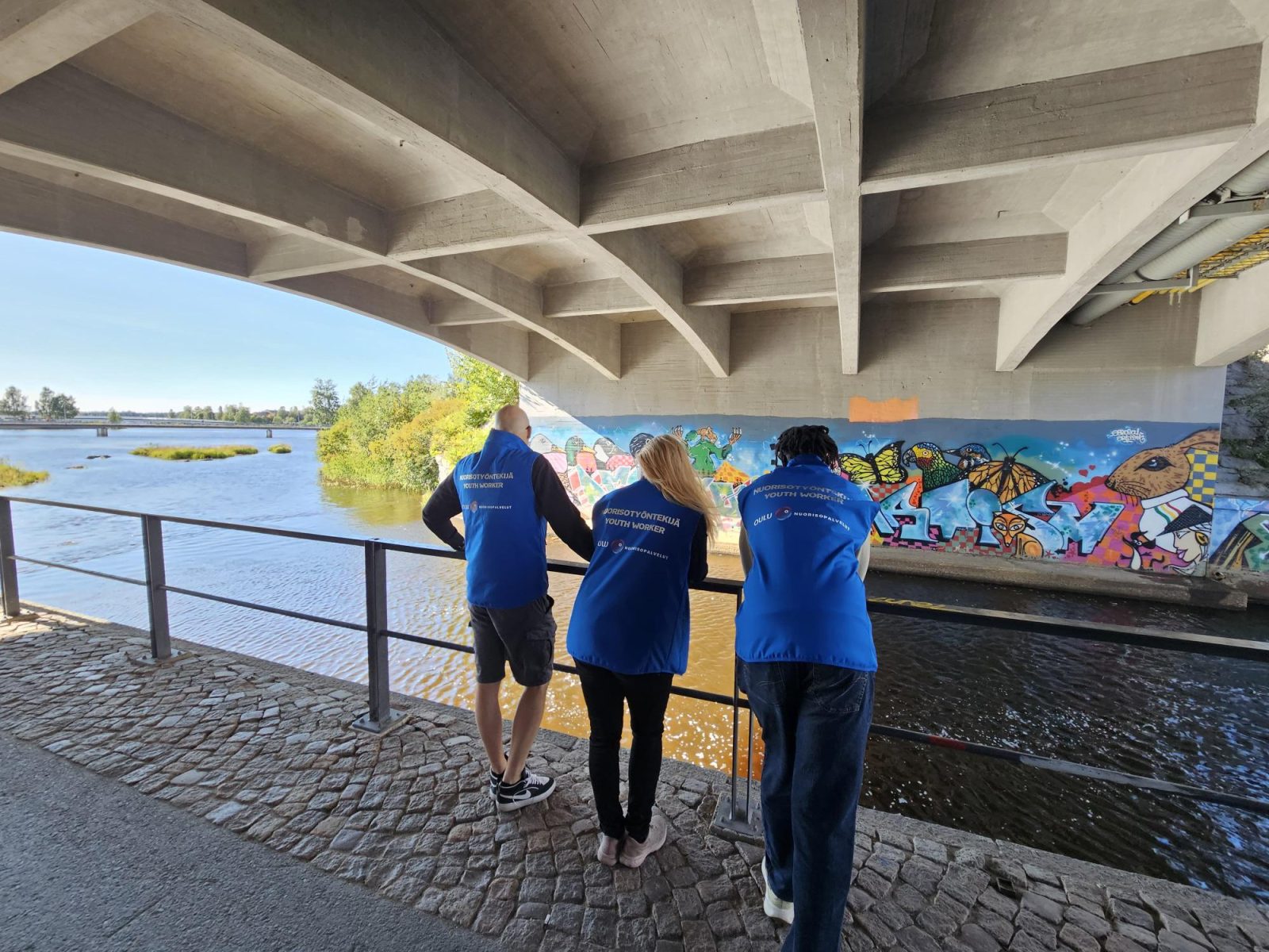 Kolme henkilöä nojaa kaiteeseen sillan allla ja taustalla näkyy Oulun suistoalue kirjastolle päin.