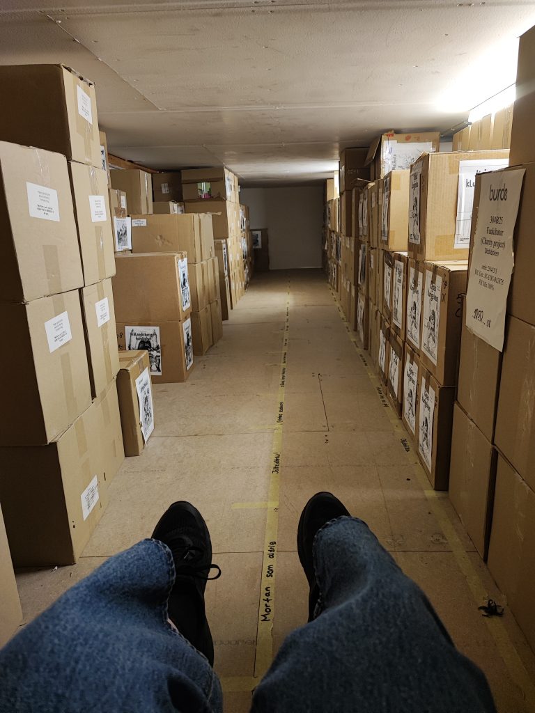 Kuvassa pahvilaatikoita kahdella seinällä kasattuna ja etualalla istuvan henkilön jalat.