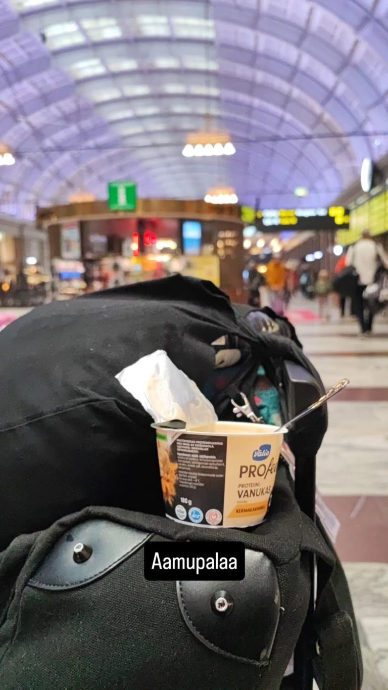 Kuvassa proteiinivanukaspurkki matkalaukun päällä. Taustalla juna-aseman infopiste. Kuvassa teksti aamupalaa.