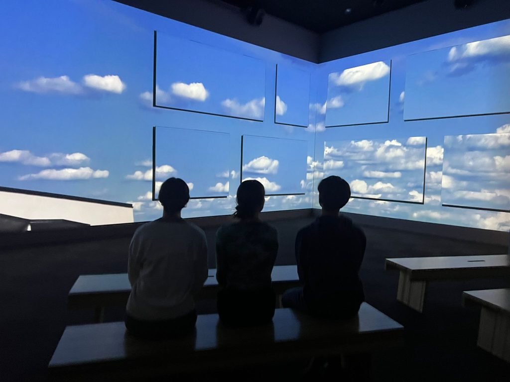 Kolme ihmistä istuu selin katsellen pilvimaisemaa.