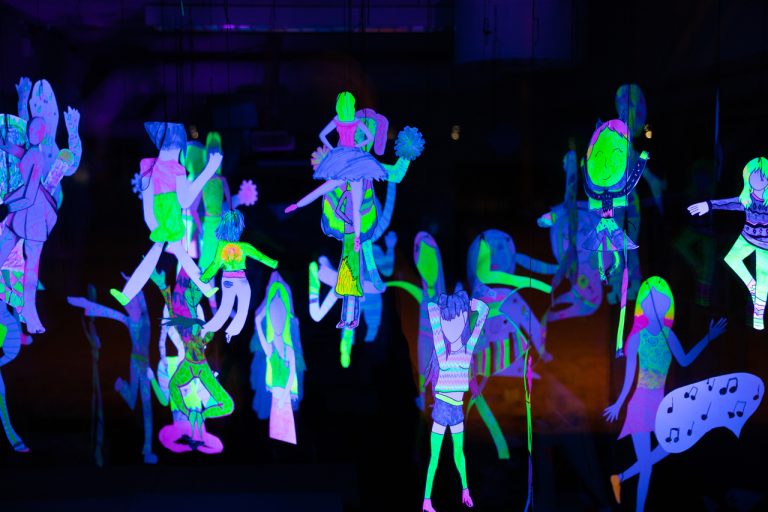 Kuvassa näkyy pimeässä hohtavia, roikkuvia lasten ja nuorten piirtämiä ihmishahmoja neon väreissä.