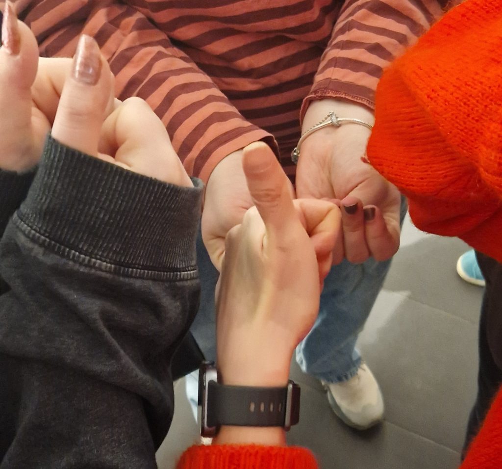 Kolme kättä, joissa peukut pystyssä, ranteessa kello. Raitapaitainen henkilö, jolla sormet supussa.