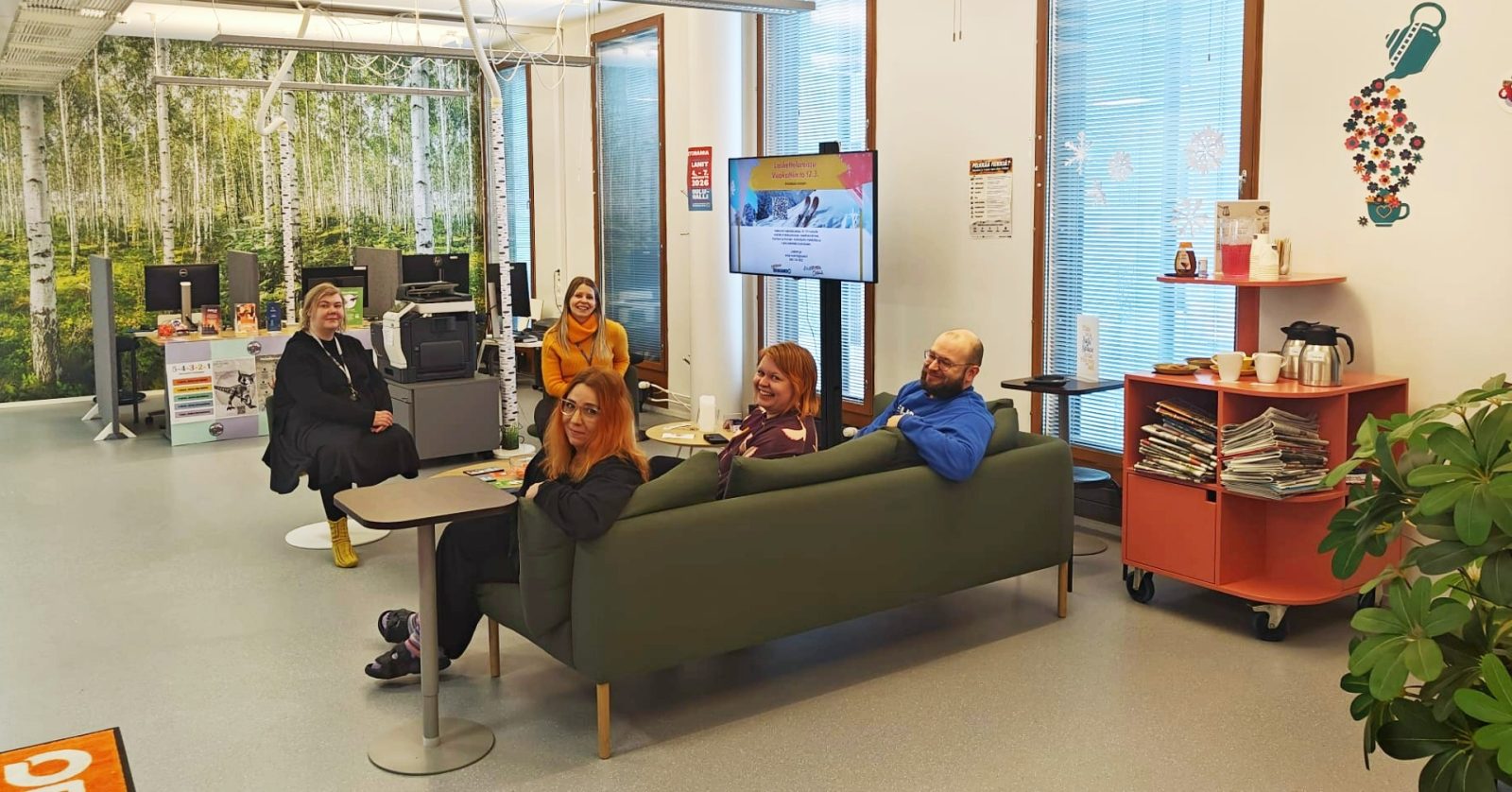 Aula, jossa viisi hymyilevää ihmistä istumassa pöydän äärellä. Taustalla koivukuosinen seinä ja neljä tietokoneen näyttöä. Neljä isoa ikkunaa sivuseinällä. Viherkasvi edustalla ja kahvipiste seinustalla.