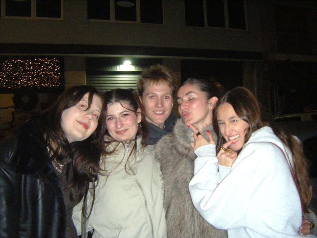 Viisi nuorta aikuista poseeraavat iloisesti iltahämärässä.