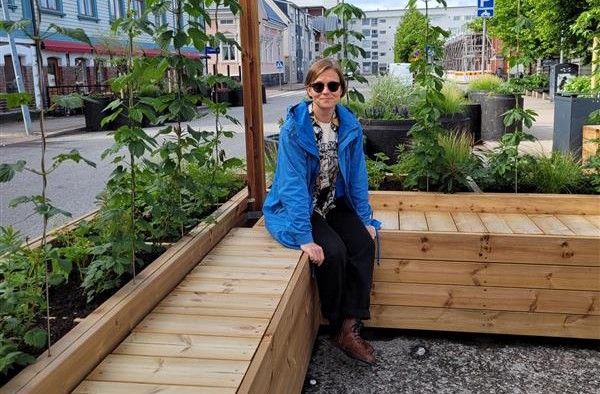 A blue-jacketed smiling adult wearing sunglasses sitting on a wooden park bench in spring weather.