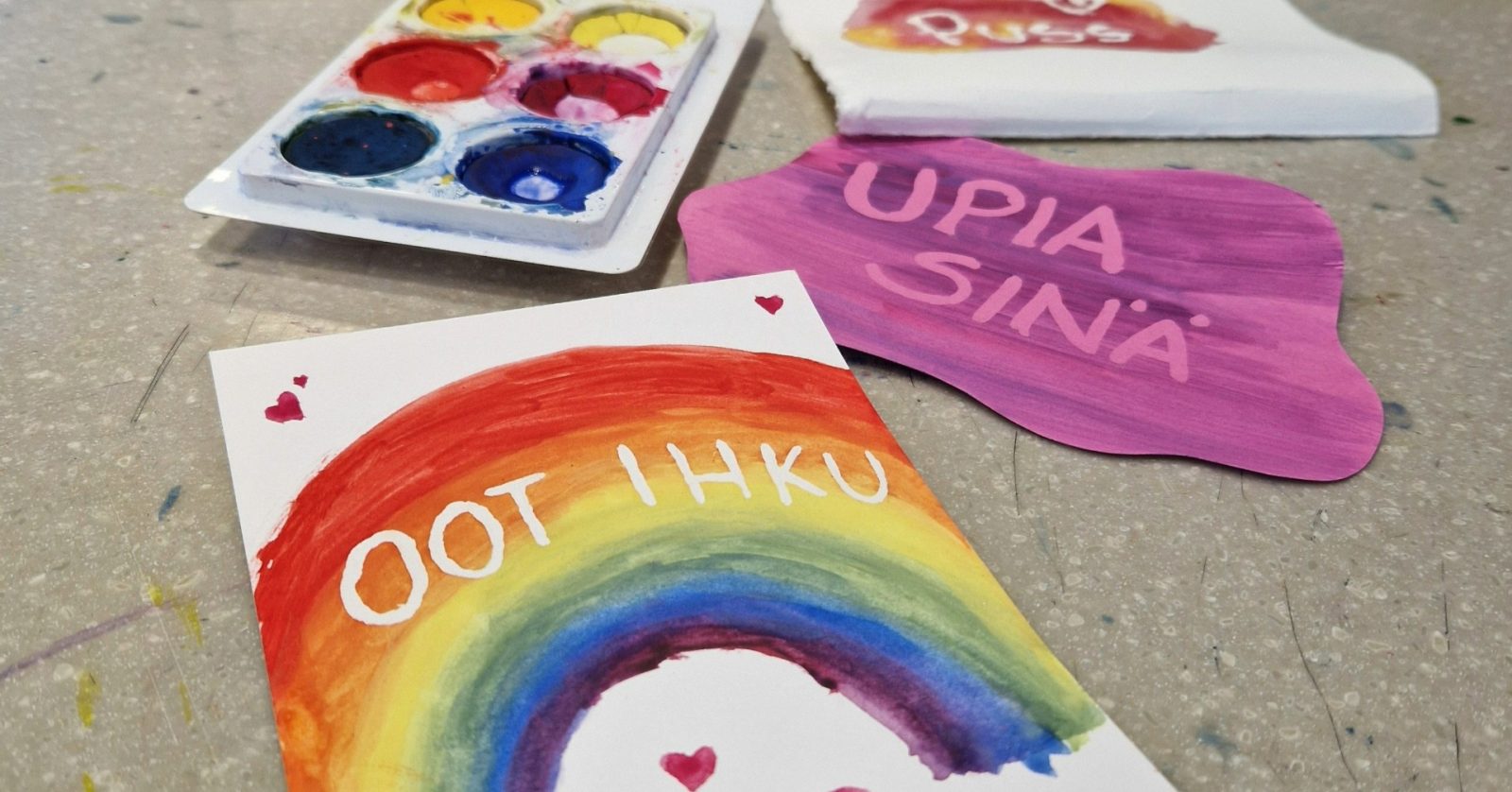 A watercolor palette and three small watercolor works. One has a rainbow with hearts and the text 'oot ihku'. One has the text 'upia sinä' and one says 'puss'.