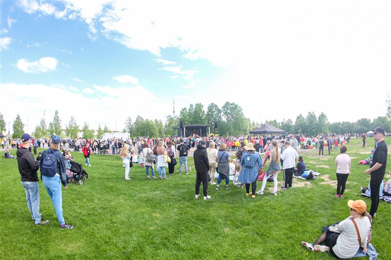 Many young people outside on the green grass. Blue sky, few clouds. Festival stage in the background.