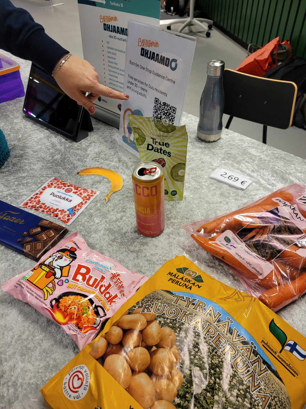Esittelypöydällä on perunapussi, porkkanapussi, energiajuomatölkki, taatelipussi, suklaalevy, Buldak -nuudelipaketti, puolukkapussin ja banaanin kuvat, sekä Byströmin Ohjaamon mainoksia.