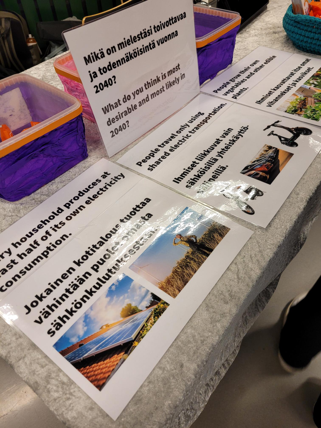 Esittelypöydällä on kolme rasiaa, joiden edessä kolme vaihtoehtoista kuvausta tulevaisuuteen liittyen. Kuvassa on myös tehtäväkortti, jossa on teksti "Mikä on mielestäsi toivottavaa ja todennäköisintä vuonna 2040?"