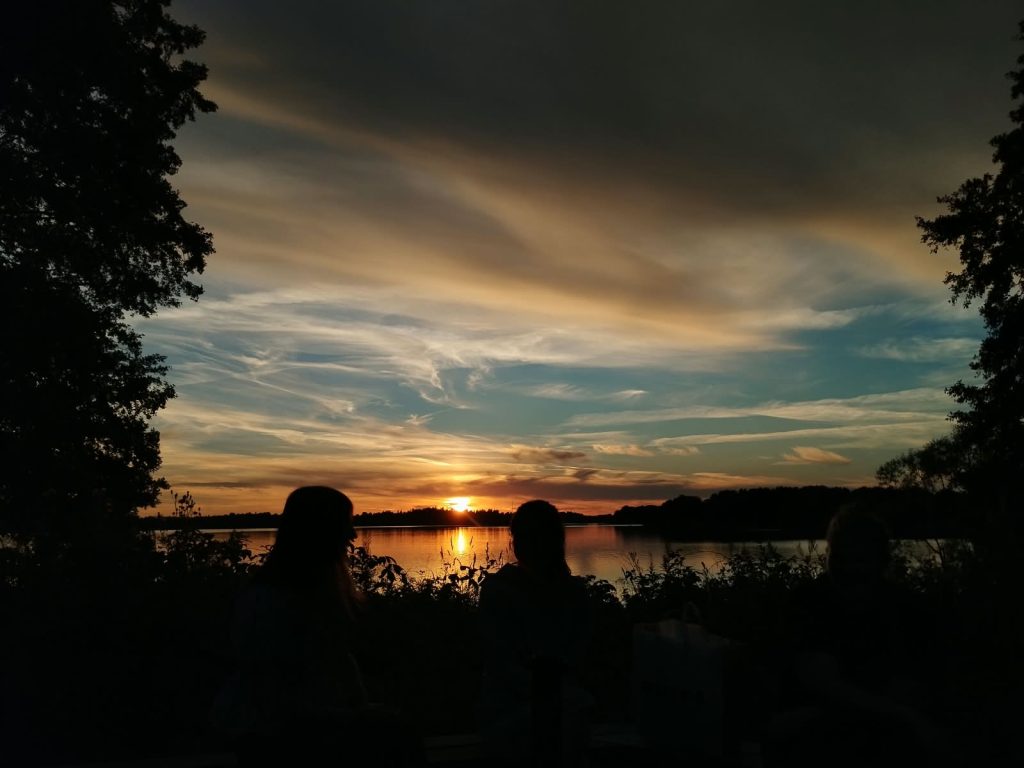 Maisemakuva auringonlaskusta järveltä ja edessä näkyy kaksi ihmishahmoa.